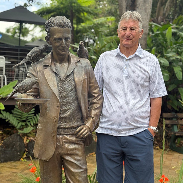 Man_in_shirt_and_shorts_standing_next_to_memorial_statue_looking_alike_surrounded_by_flowers_and_trees.jpg
