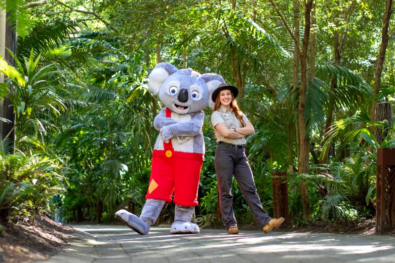 A person in a life-size Koala costume with red pants and a woman in kaki outdoor clothing, boots and a stetson hat, posing on a stone footpath surrounded by green tropical trees and bushes
