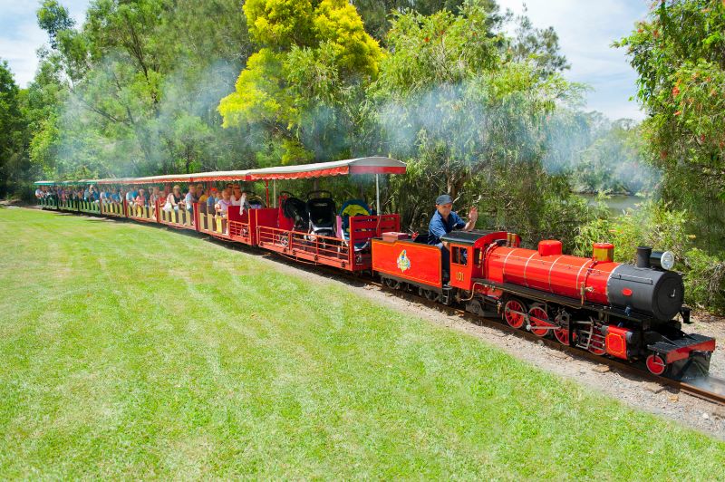 Red mini train carrying passengers through a park, passing green grass large trees and a water pond