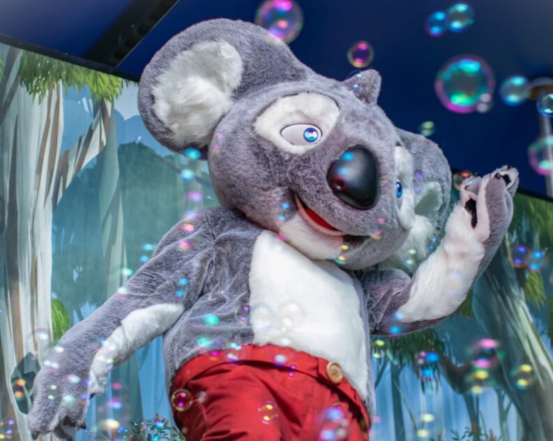 A person in a koala costume with red pants waves while surrounded by floating bubbles, with a forest-themed backdrop behind.