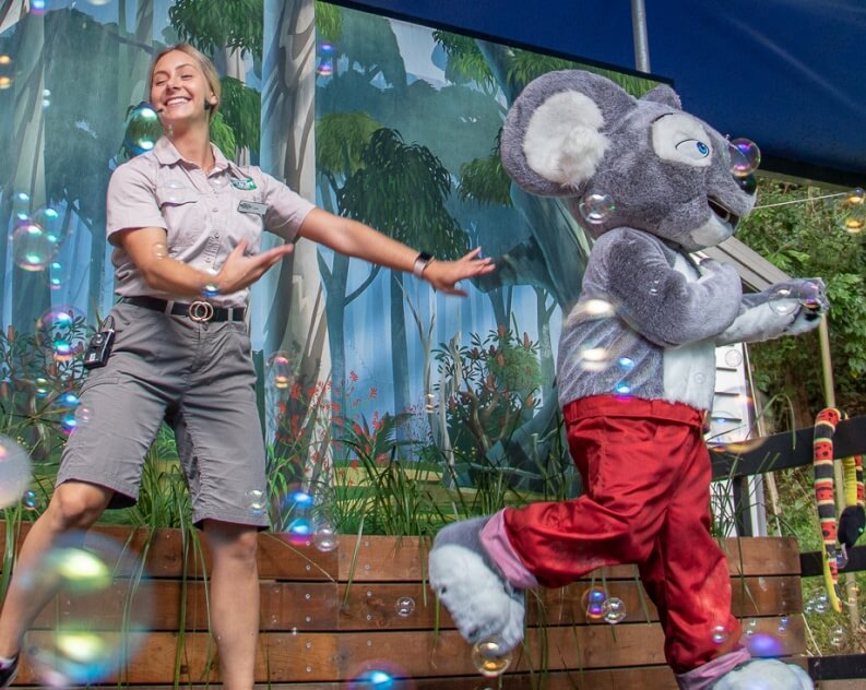 A woman in uniform and a person in a koala costume perform amid floating bubbles on an outdoor stage with a forest backdrop.