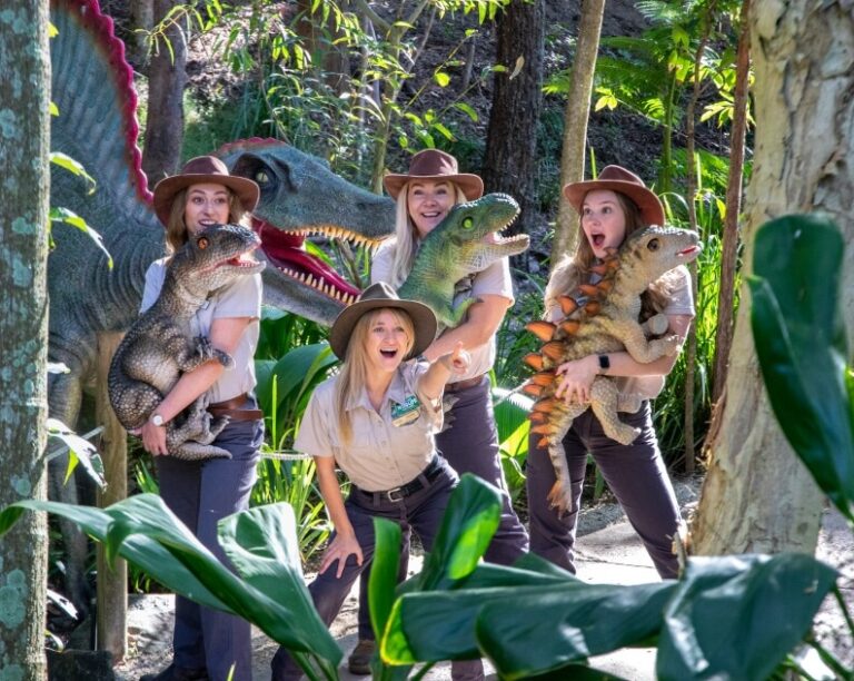 Four people in ranger uniforms pose in a forest setting with toy dinosaurs and a large dinosaur statue behind them.