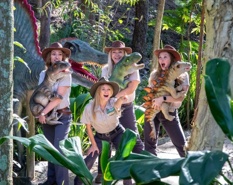 Four people in ranger uniforms pose in a forest setting with toy dinosaurs and a large dinosaur statue behind them.
