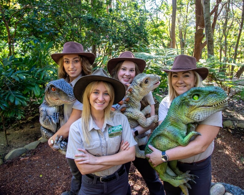 Four women in khaki uniforms and brown hats pose outdoors, each holding a realistic dinosaur puppet, surrounded by lush greenery.