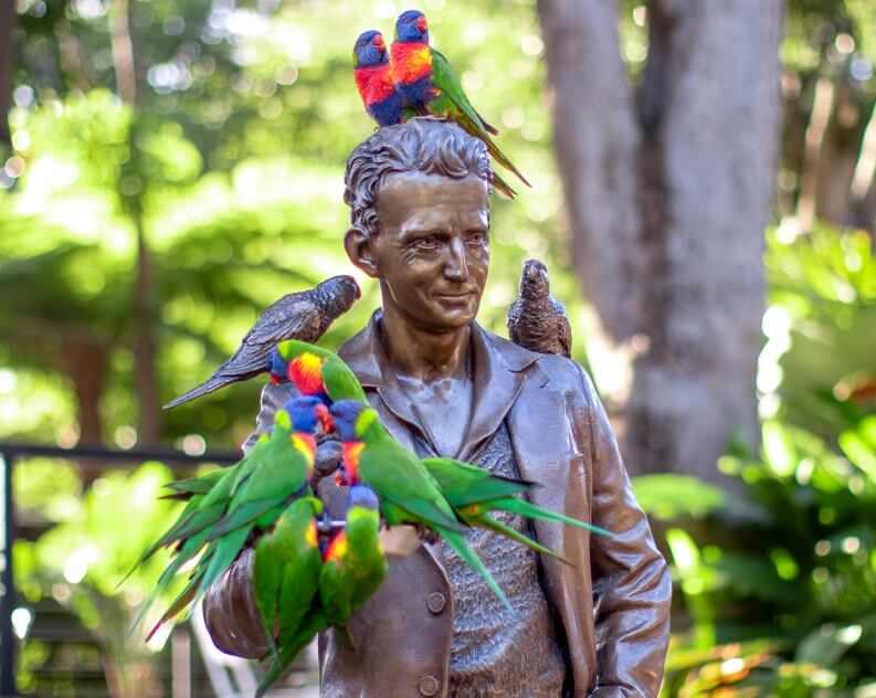 Bronze statue of a man surrounded by several colorful parrots perched on his head, shoulders, and arm, with greenery in the background.