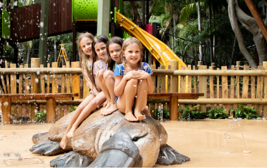 Four young girls sitting on a large platypus sculpture in a water play area, smiling and enjoying the outdoor setting, with slides and greenery in the background