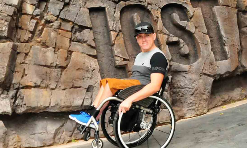 Person in a wheelchair smiling in front of a rock wall with large engraved letters.