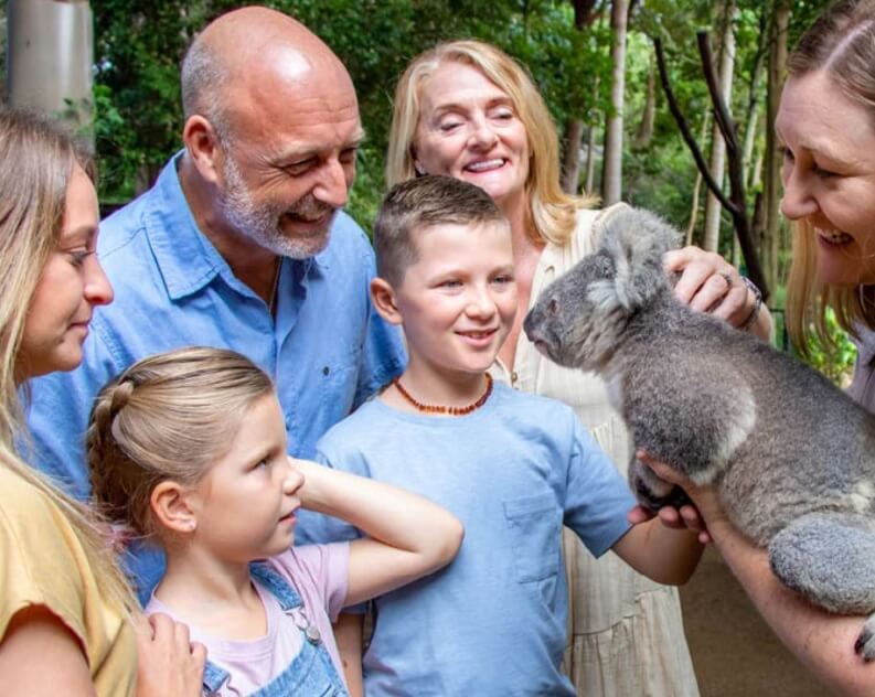 A family gathers around a zookeeper holding a koala, smiling and looking at the animal in an outdoor setting.