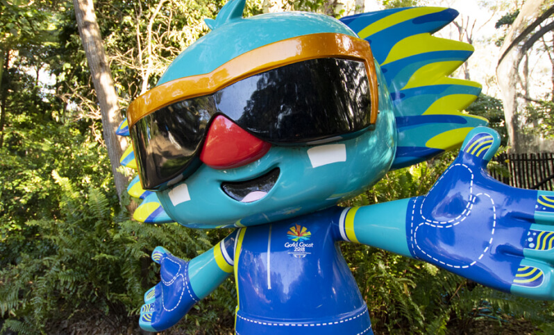 Large, teal mascot figure with sunglasses, a red nose, and a Gold Coast logo, standing amidst greenery.
