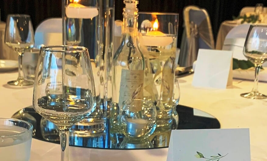 Table setting with clear glass vases, candles, wine glasses, and place cards at a wedding reception.