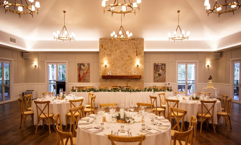 Wedding reception hall with round tables, white linens, and light wooden chairs.