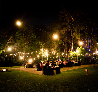 Outdoor nighttime event with string lights, tables, and people gathered.