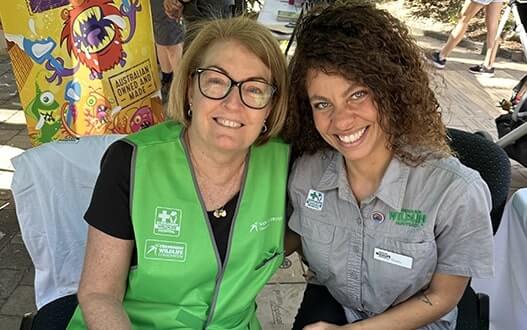 Two women in green and gray wildlife conservation vests smile at the camera, standing near a colorful Australian-made children's book display.