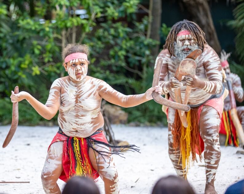 Two Indigenous Australian performers wearing traditional attire and body paint perform a cultural dance outdoors, holding boomerangs.