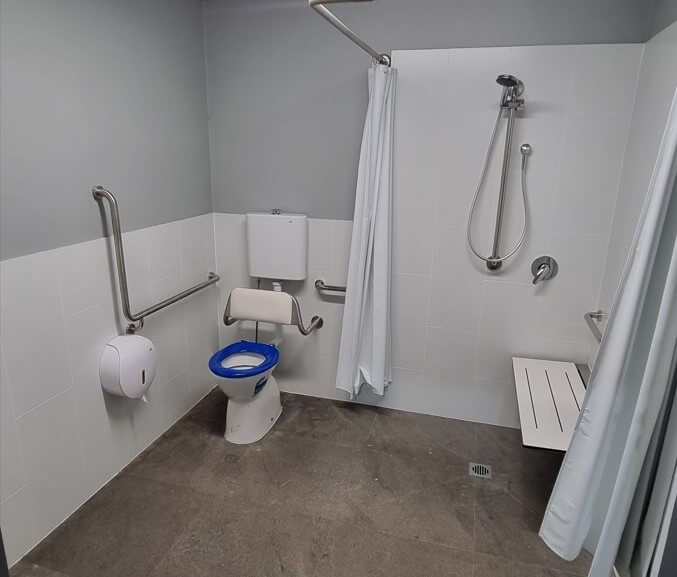 A well-equipped accessible bathroom featuring a toilet with a blue seat and backrest, grab bars for support, a shower area with a handheld showerhead, a shower curtain, and a foldable shower seat.