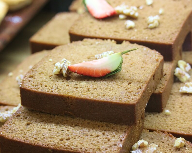 Several slices of dense, moist brown bread stacked together, topped with small walnut pieces and a colorful red and green garnish, possibly a strawberry or decorative fruit.