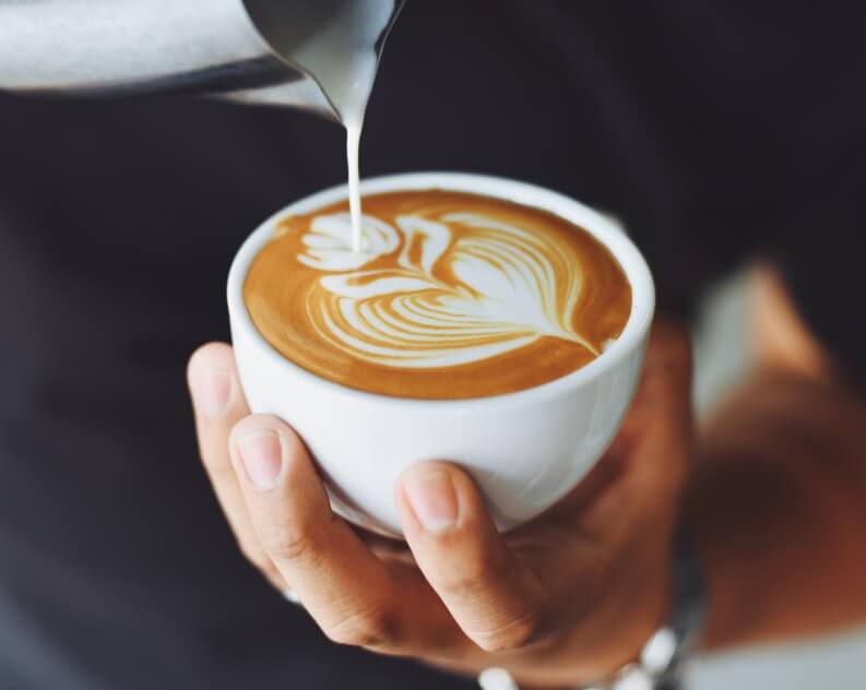 A close-up image of a hand holding a white coffee cup, filled with coffee featuring an intricate floral latte art pattern.