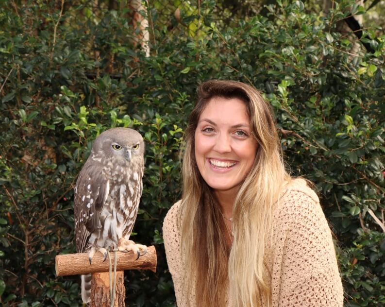 A woman with long blonde hair smiles next to a grey and white owl perched on a wooden stand, with green foliage in the background.