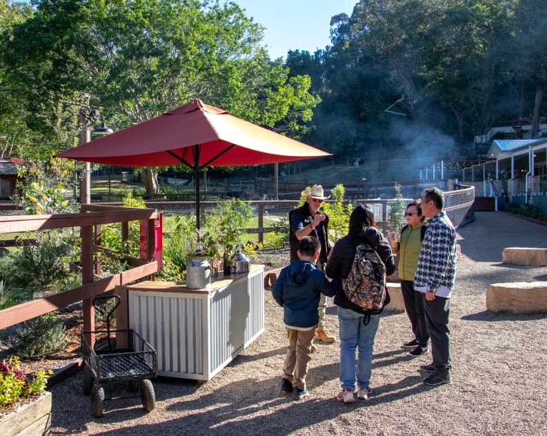 A group of people stands near an outdoor kiosk with a red umbrella, listening to a person in a hat; trees and fencing appear in the background.