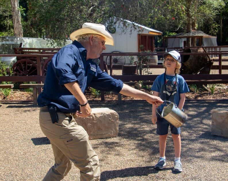 An adult in a hat pours water from a kettle while a child watches outside in a sunny, rustic setting.