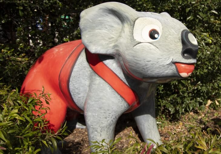 A large, painted koala statue wearing red suspenders stands among green bushes and foliage.