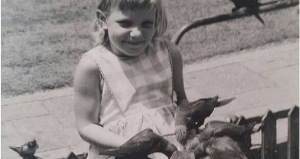 A black and white photograph of a young child wearing a sleeveless checkered dress, standing on a paved area outdoors with grass in the background.