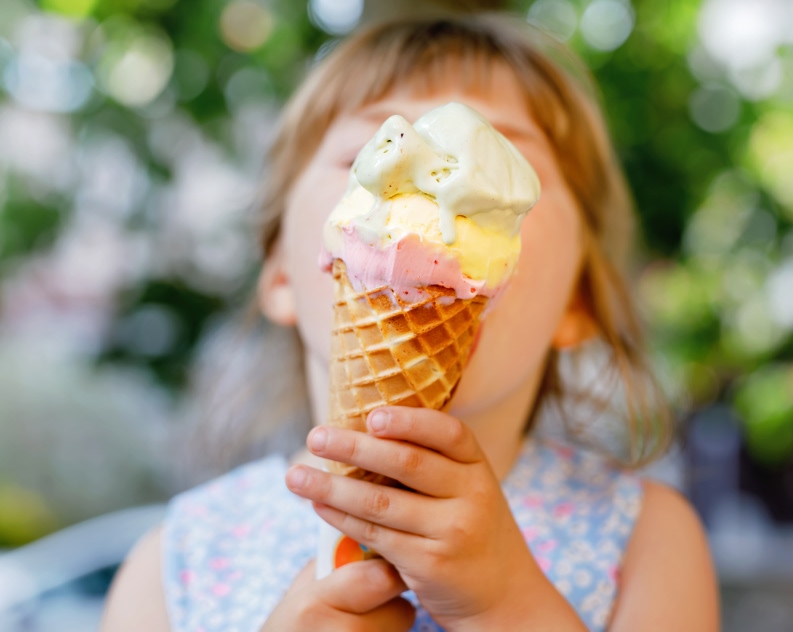 A young child wearing a sleeveless floral dress holds a single-scoop ice cream cone. The background features greenery and blurred elements, suggesting a park or garden environment.