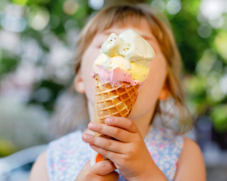 A young child in a sleeveless floral dress holding an ice cream cone with both hands. The background is softly blurred, showcasing lush greenery, suggesting an outdoor setting.