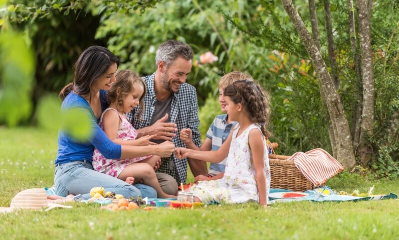 A group of people sitting on a picnic blanket in a grassy area surrounded by lush greenery and trees.