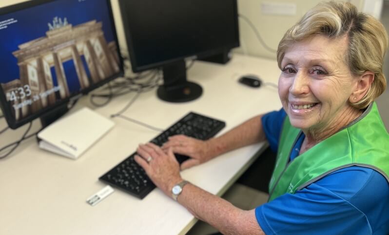 Older woman wearing a green and blue shirt smiles at the camera while sitting at a desk and using a computer.
