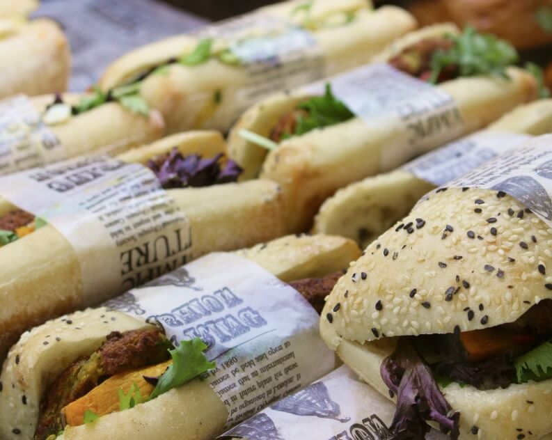 A variety of sandwiches wrapped in paper, featuring different types of bread such as plain and sesame seed varieties.