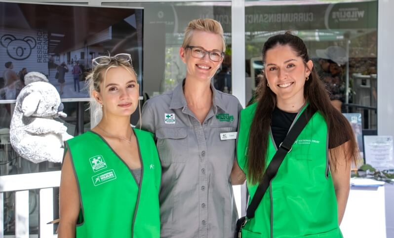 Three women wearing uniforms stand and smile at an indoor event; two wear green vests, one wears a gray shirt. A koala image and informational displays are visible in the background.