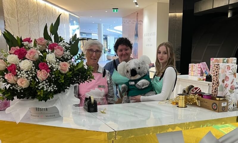 Three people stand behind a white counter with a large bouquet of flowers and gift boxes; one person holds a plush koala toy.