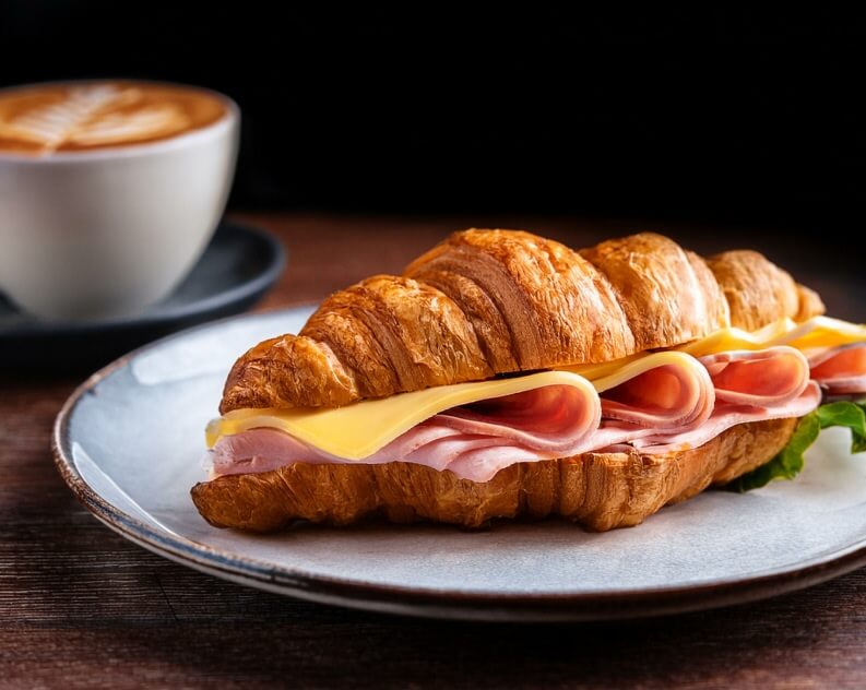 A flaky croissant sandwich, filled with layers of sliced ham and melted cheese, placed on a plate. A small piece of lettuce peeks from the sandwich, adding a fresh touch.