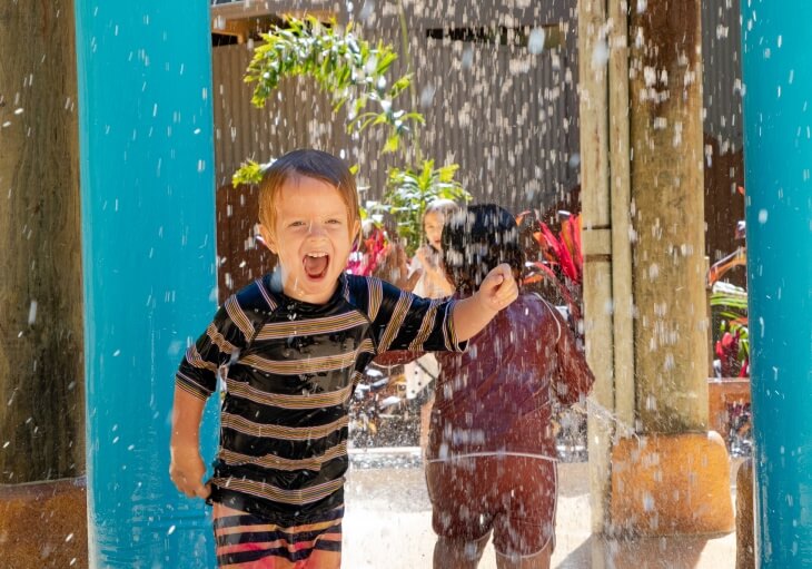 Young child in striped swimwear smiles and runs through water spray at an outdoor play area, with another child standing in the background.