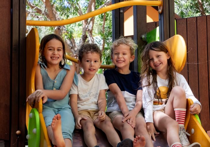 Four children sit side by side at the top of a playground slide, smiling at the camera with trees and wooden structures in the background.