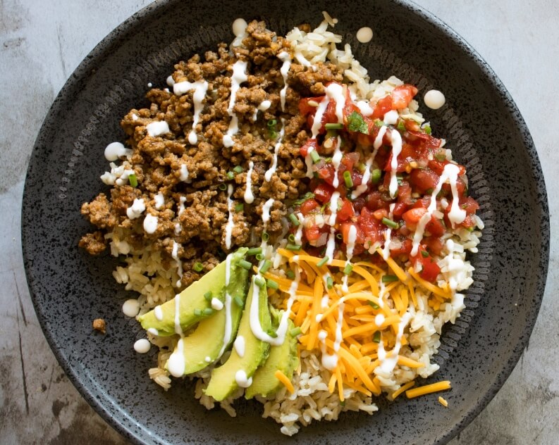 A vibrant burrito bowl filled with seasoned ground meat, fluffy rice, diced tomatoes with herbs, shredded cheese, sliced avocado, and a drizzle of creamy white sauce.