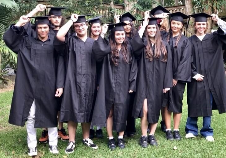 A group of nine graduates wearing black caps and gowns stand on grass outdoors, smiling and holding their caps.