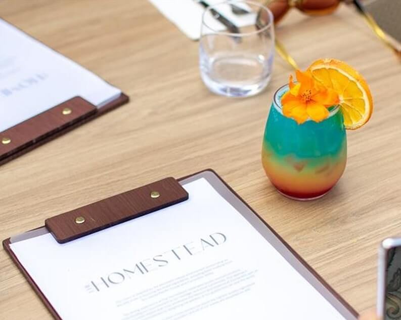 Wooden-handled menu clipboard sits beside a rainbow layered cocktail garnished with orange slice and flower.