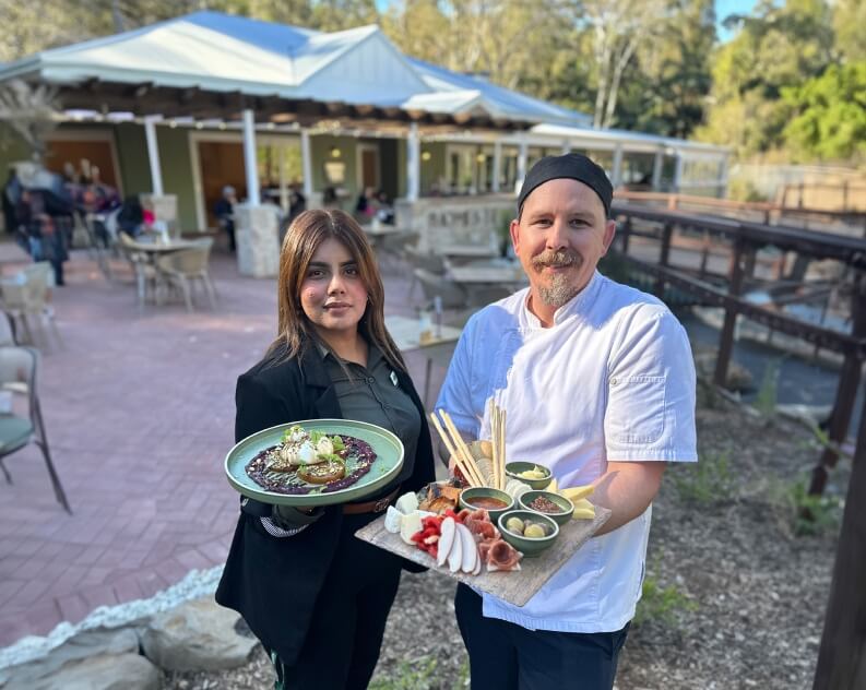 Two people, one in a chef's uniform and the other in a blazer, hold platters of food outdoors at a restaurant.