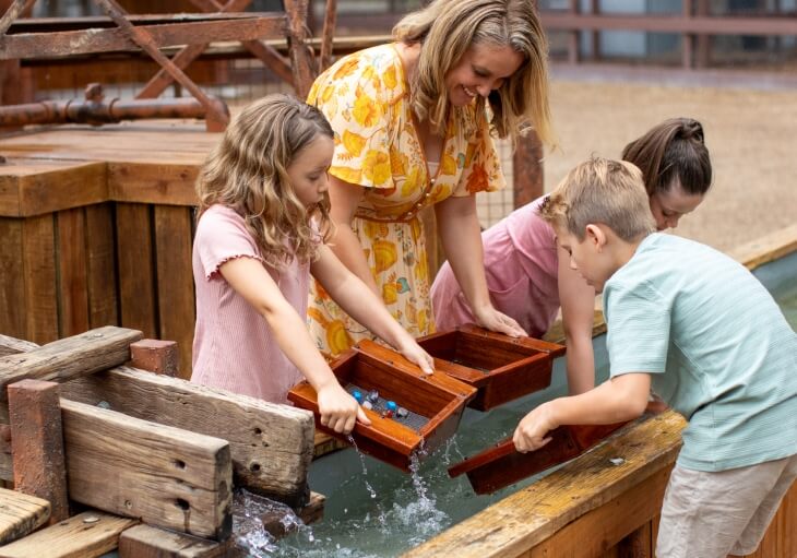 An adult and three children use wooden pans to sift through water at an outdoor activity station.