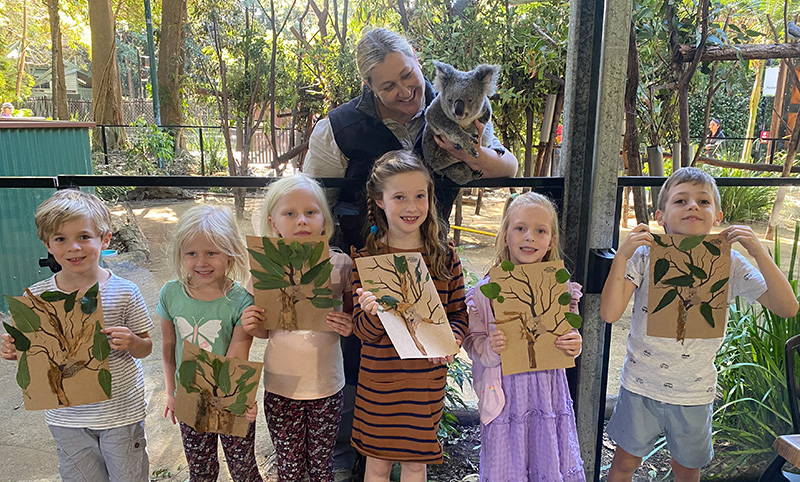 Group of young kids holding up drawing. Wildlife keeper and koala standing behind them