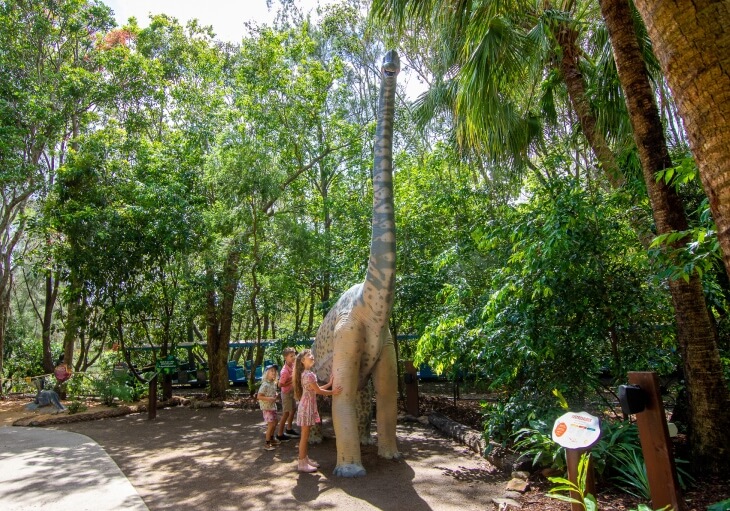 Children interact with a life-sized Brachiosaurus statue surrounded by lush greenery on a sunny day in a park or zoo.