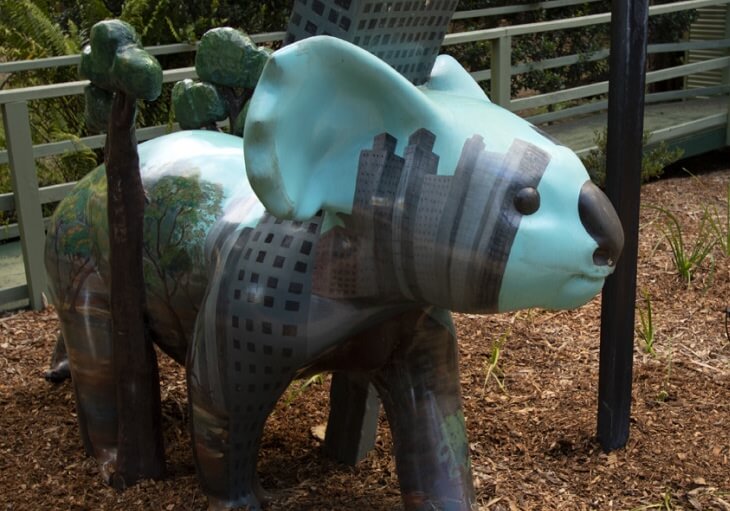 A painted koala statue featuring city buildings and trees on its body stands outdoors on wood chips near a railing.
