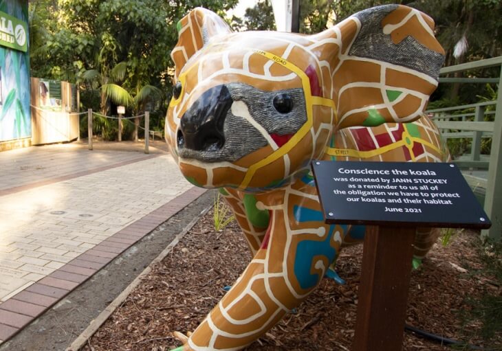 A colorful koala statue with a patterned design stands beside a sign explaining it was donated as a reminder to protect koalas and their habitat. Trees and a walkway are visible in the background.