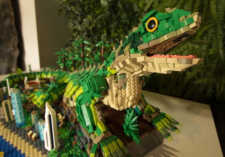 A large green lizard sculpture made from LEGO bricks is displayed on a table, surrounded by small LEGO trees and buildings.