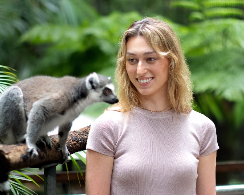 A woman standing next to a ring-tailed lemur on a branch, surrounded by greenery.