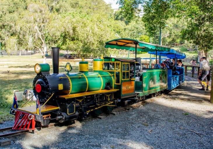 A small green and yellow open-air train with several passengers travels along a shaded track in a park setting.
