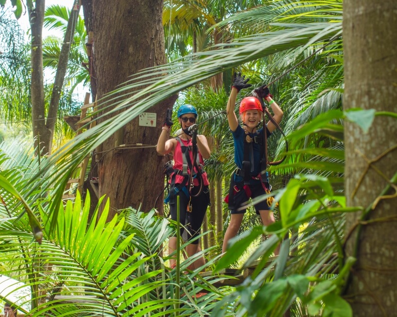 Two people wearing helmets and harnesses stand on a platform in a tropical forest, preparing for a zipline adventure among lush green foliage.