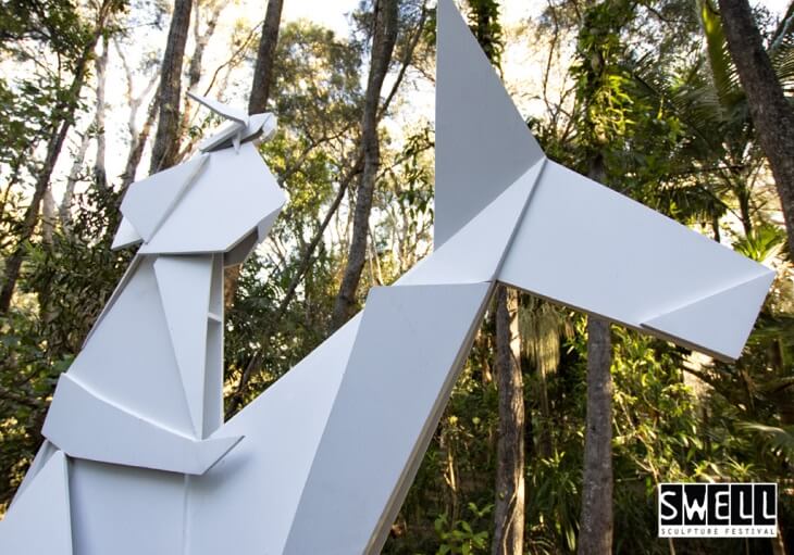 Large white geometric horse sculpture set among tall trees at an outdoor location, with SWELL Sculpture Festival logo in the corner.